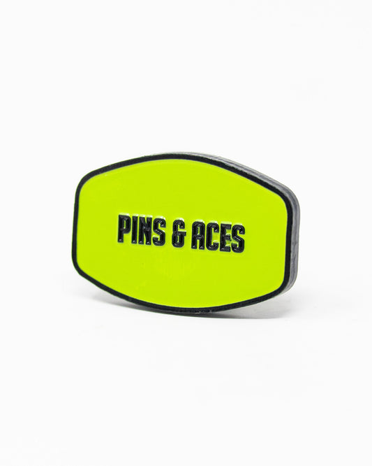 A yellow-green HEYDUDE - Logo Ball Marker by Pins and Aces, with black edges and bold black "PINS & ACES" text on the front, set against a plain white background.