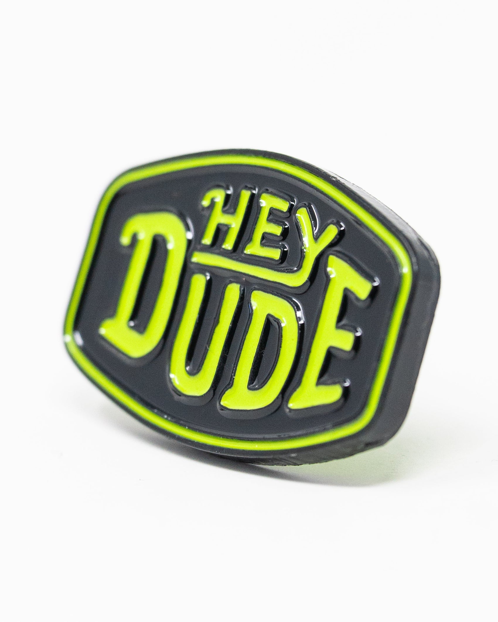 The Pins and Aces HEYDUDE - Logo Ball Marker is a black cast metal marker with rounded edges, featuring bold raised yellow-green "HEY DUDE" text outlined in the same color. Shown on a white background.
