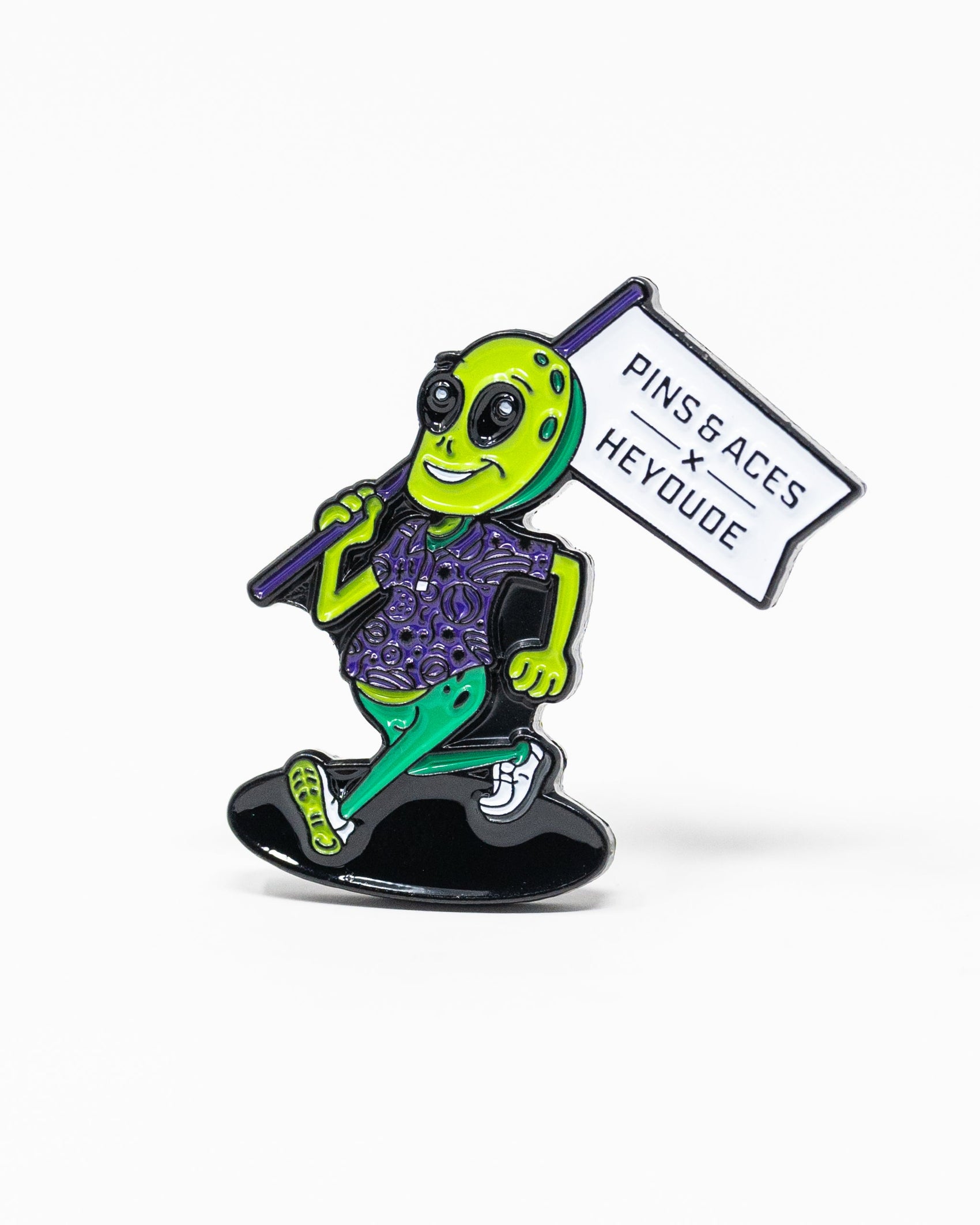 Show off your style with the Pins and Aces HEYDUDE – Alien Ball Marker: a vibrant enamel pin of a smiling green alien in a purple shirt, green sandals, and holding a PINS & ACES X HEY DUDE sign.