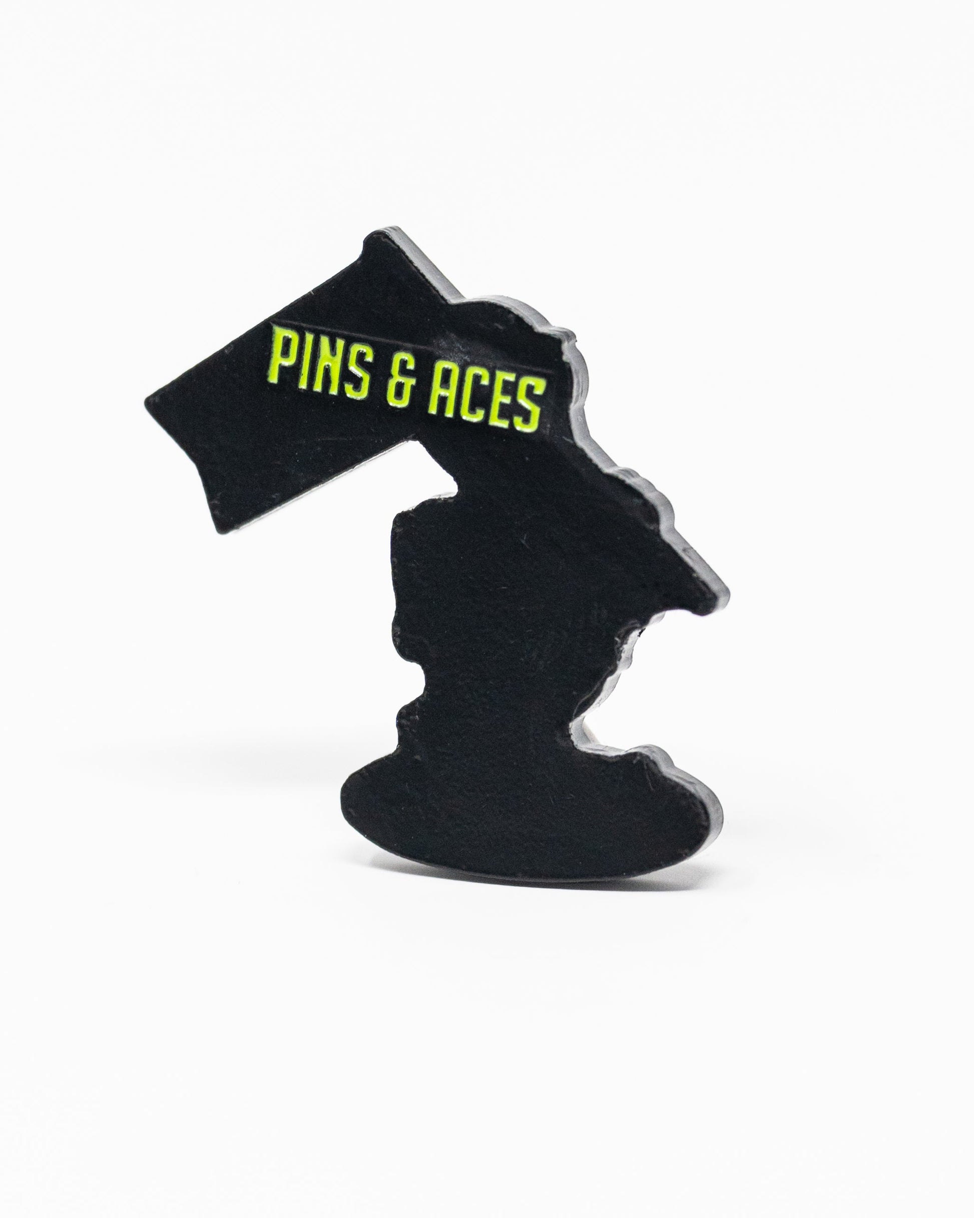 A black enamel HEYDUDE - Alien Ball Marker by Pins and Aces, shaped like Michigan, features bold yellow branding on a flag and is photographed against a plain white background.