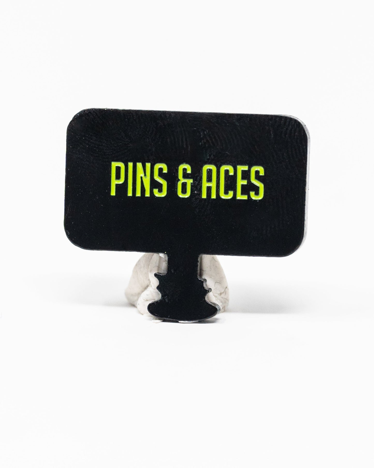 A rectangular black sign with yellow-green “PINS & ACES” text stands upright on gray clay—ideal for displaying the Pins and Aces HEYDUDE - Hole 51 Ball Marker by Pins and Aces.