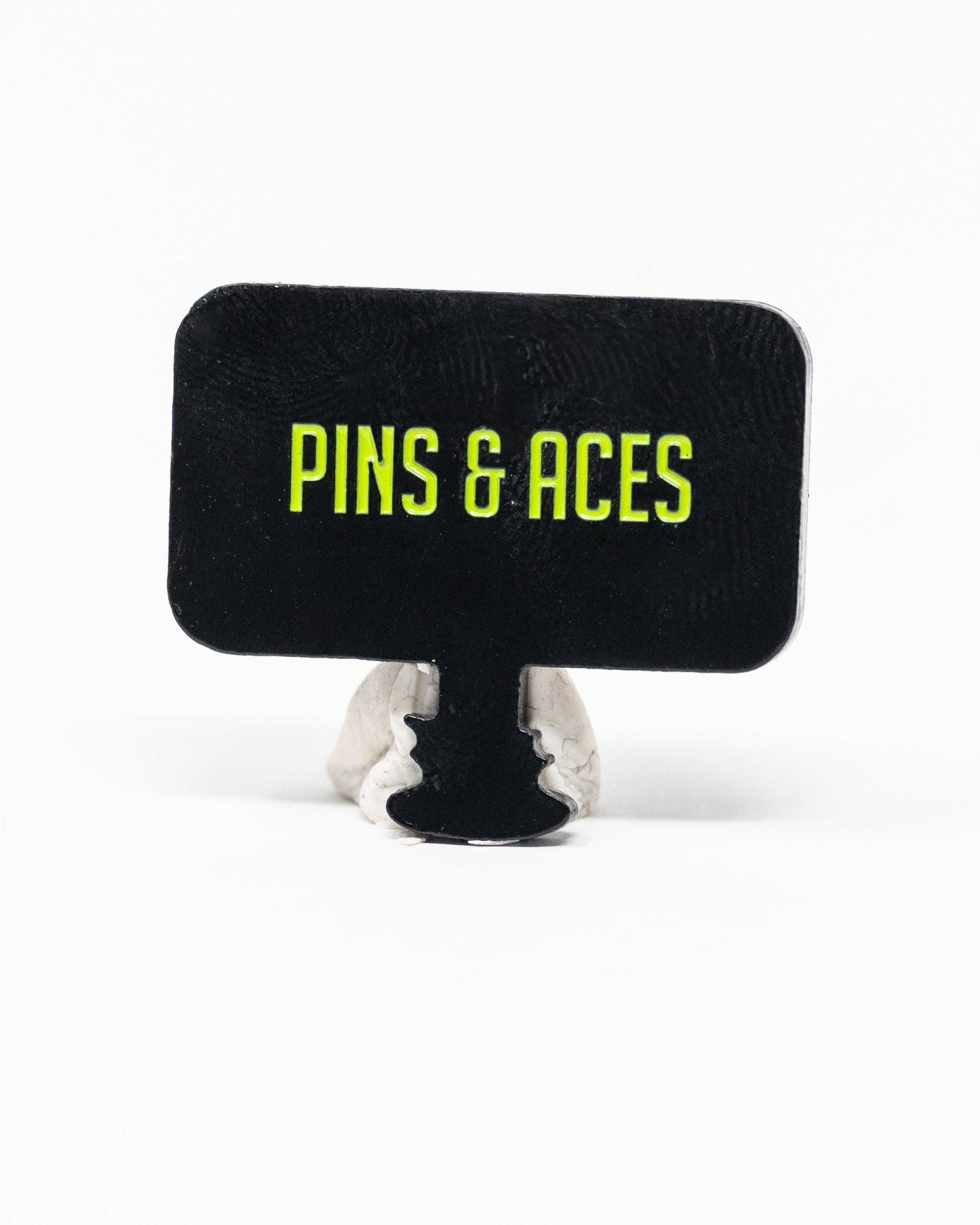 A rectangular black sign with yellow-green “PINS & ACES” text stands upright on gray clay—ideal for displaying the Pins and Aces HEYDUDE - Hole 51 Ball Marker by Pins and Aces.