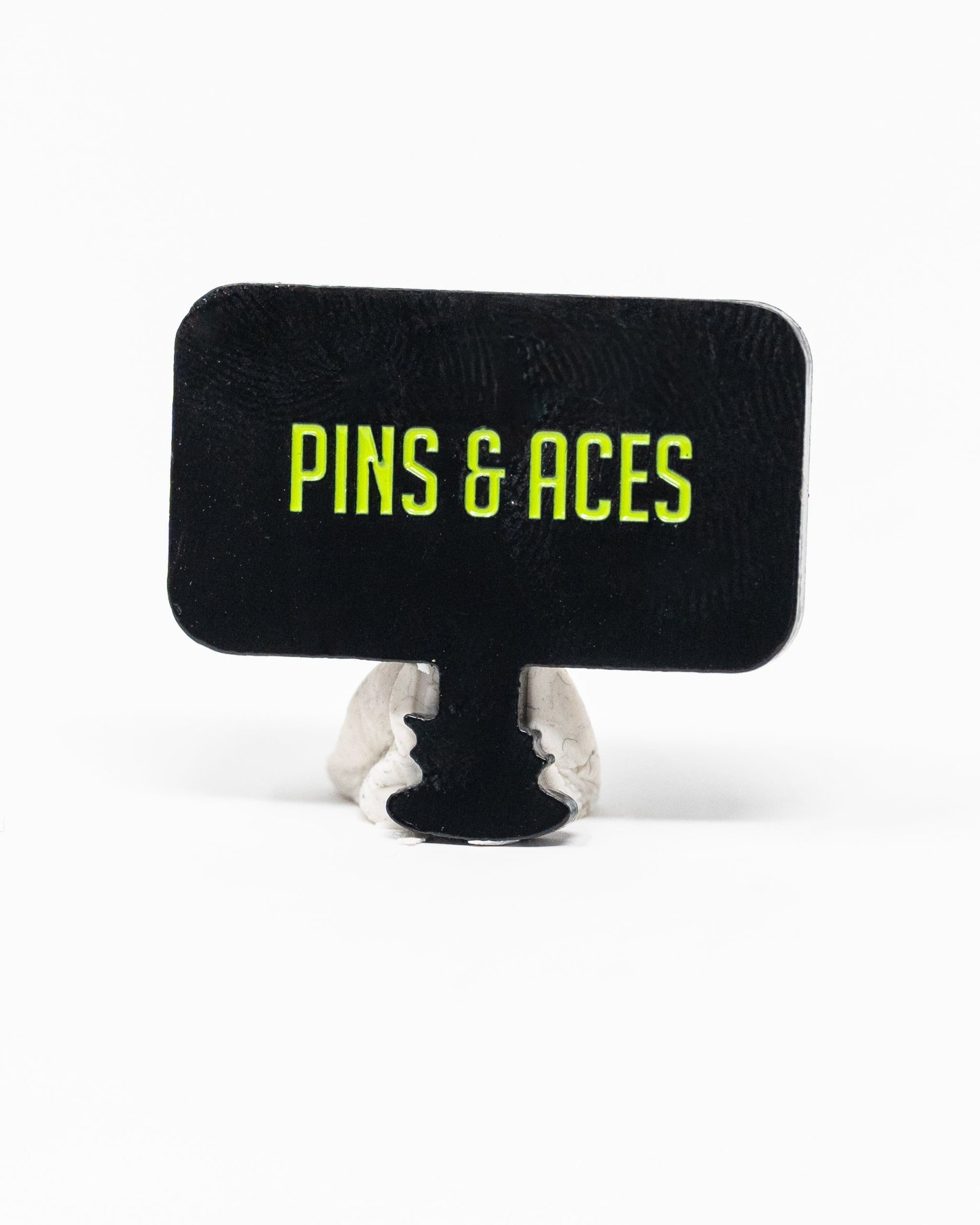 A rectangular black sign with yellow-green “PINS & ACES” text stands upright on gray clay—ideal for displaying the Pins and Aces HEYDUDE - Hole 51 Ball Marker by Pins and Aces.