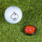 A white golf ball with a black spade and a Pins and Aces Pop Out Medallion Ball Marker - Red rest on green artificial turf.