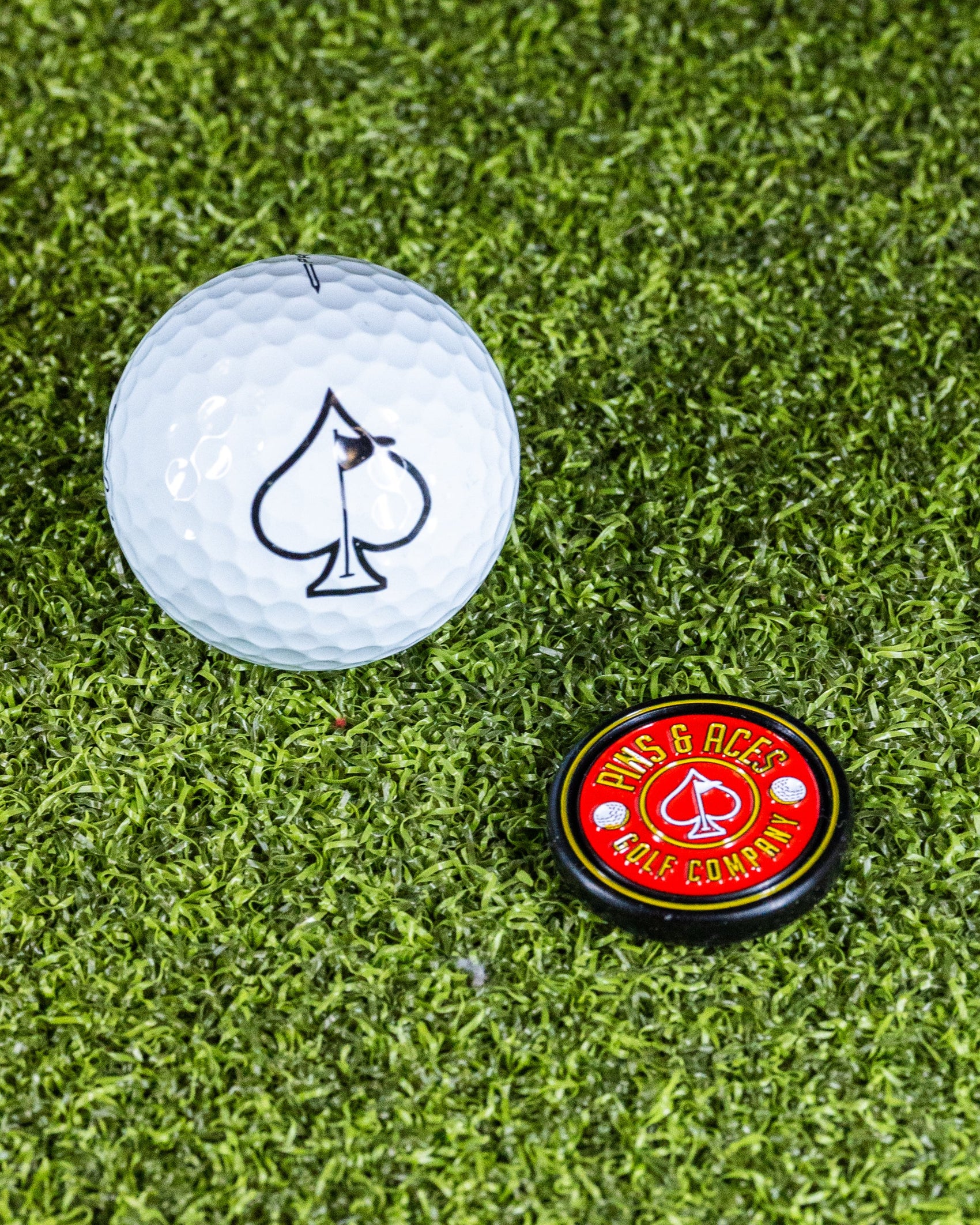 A white golf ball with a black spade and a Pins and Aces Pop Out Medallion Ball Marker - Red rest on green artificial turf.