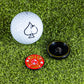 A golf ball with a spade symbol sits on grass next to a red Pop Out Medallion Ball Marker by Pins and Aces and a black magnetic holder.