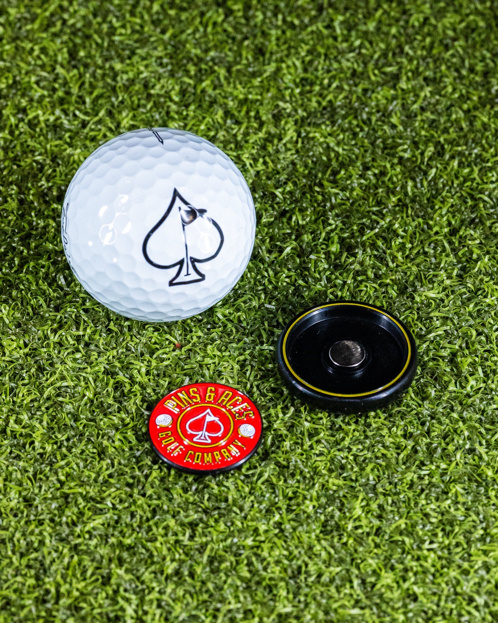 A golf ball with a spade symbol sits on grass next to a red Pop Out Medallion Ball Marker by Pins and Aces and a black magnetic holder.