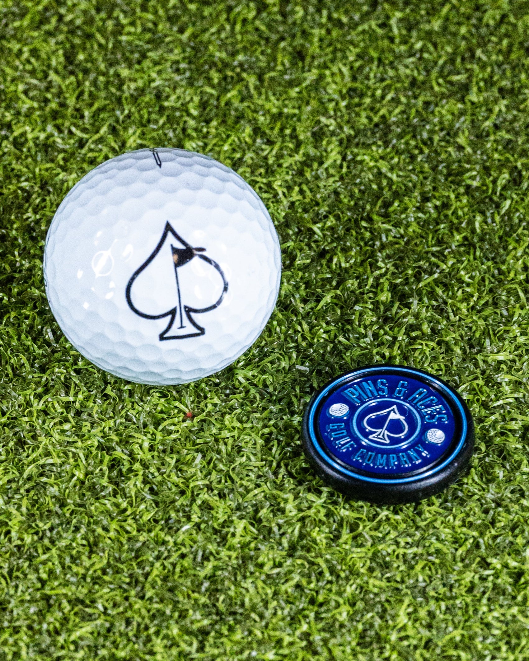 A Pop Out Medallion Ball Marker - Navy by Pins and Aces and a white golf ball with a black spade symbol rest on green artificial turf.