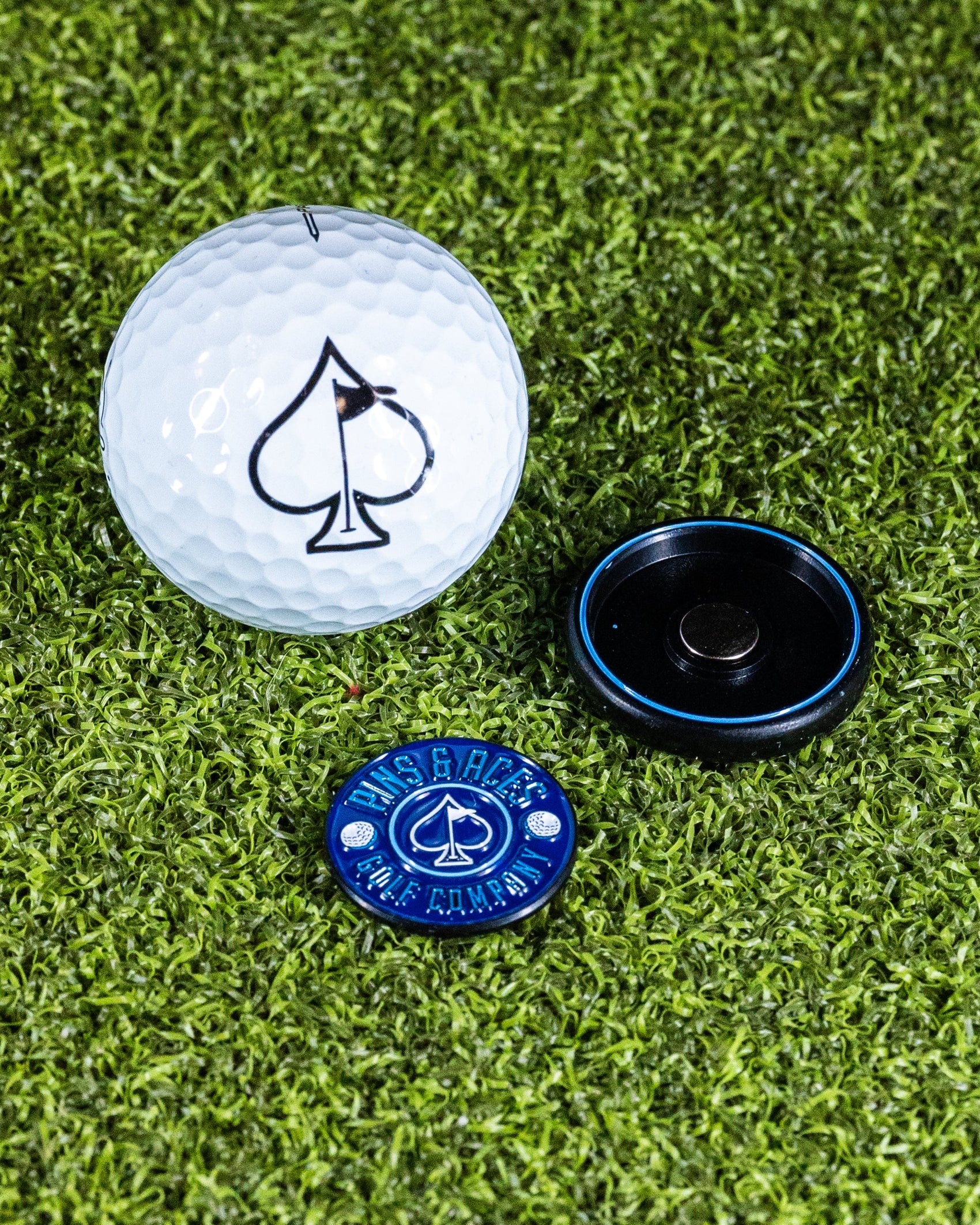 The Pins and Aces Pop Out Medallion Ball Marker - Navy, featuring a spade symbol and navy design, and a black magnetic holder are displayed on green artificial turf.