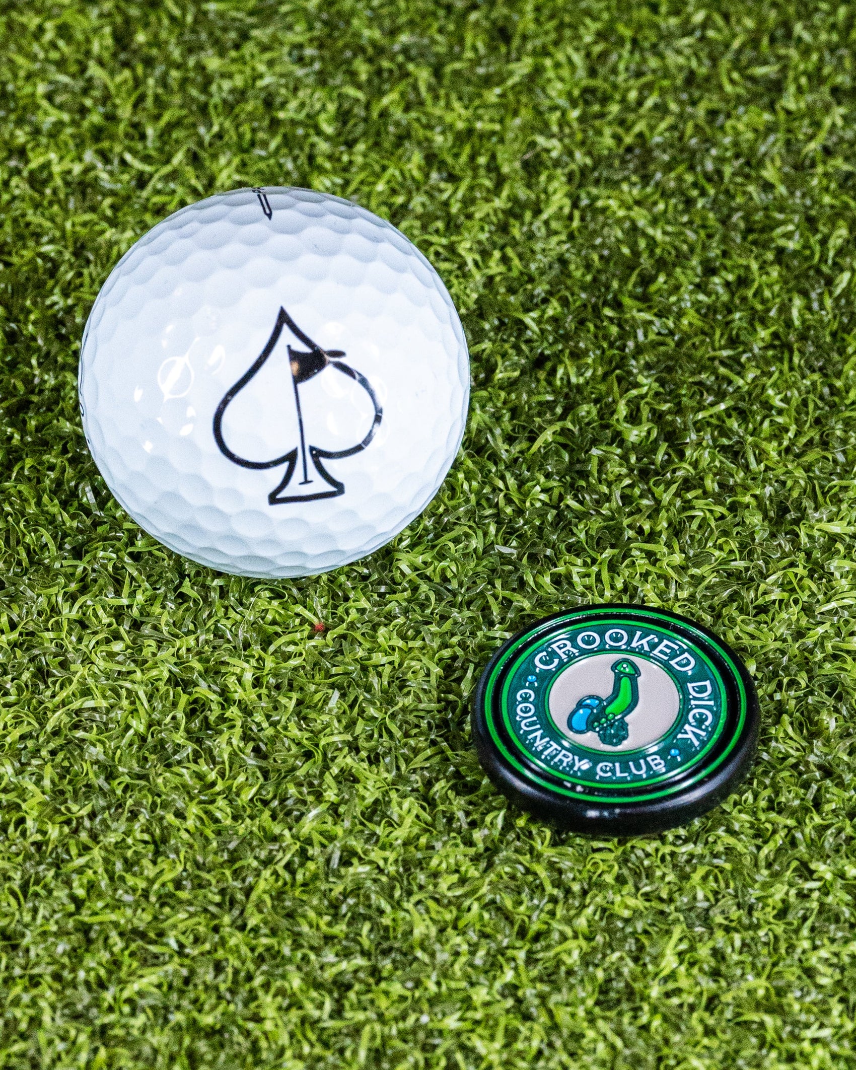 A white golf ball with a black spade and a round green Pop Out Medallion Ball Marker - Crooked Dick by Pins and Aces, featuring a bird logo, rest on green artificial turf.