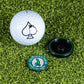 A Pins and Aces Pop Out Medallion Ball Marker - Crooked Dick, featuring a green “Crooked Dick Country Club” design and a black pop-out medallion, is displayed on artificial grass.