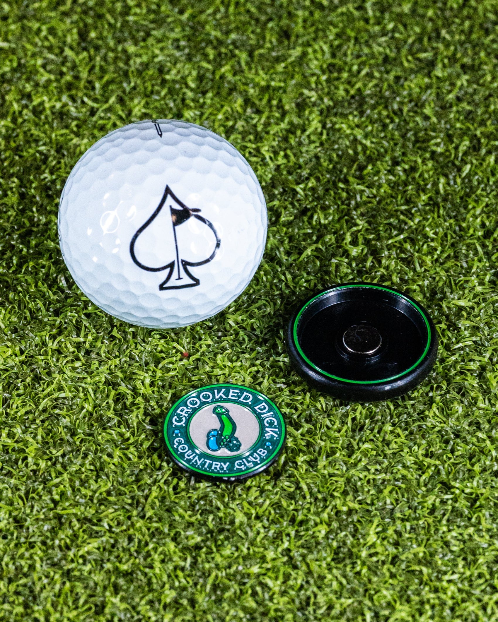 A Pins and Aces Pop Out Medallion Ball Marker - Crooked Dick, featuring a green “Crooked Dick Country Club” design and a black pop-out medallion, is displayed on artificial grass.