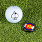 A white golf ball with a black spade symbol sits next to the Pins and Aces Pop Out Medallion Ball Marker - Colorado, both resting on green artificial turf.