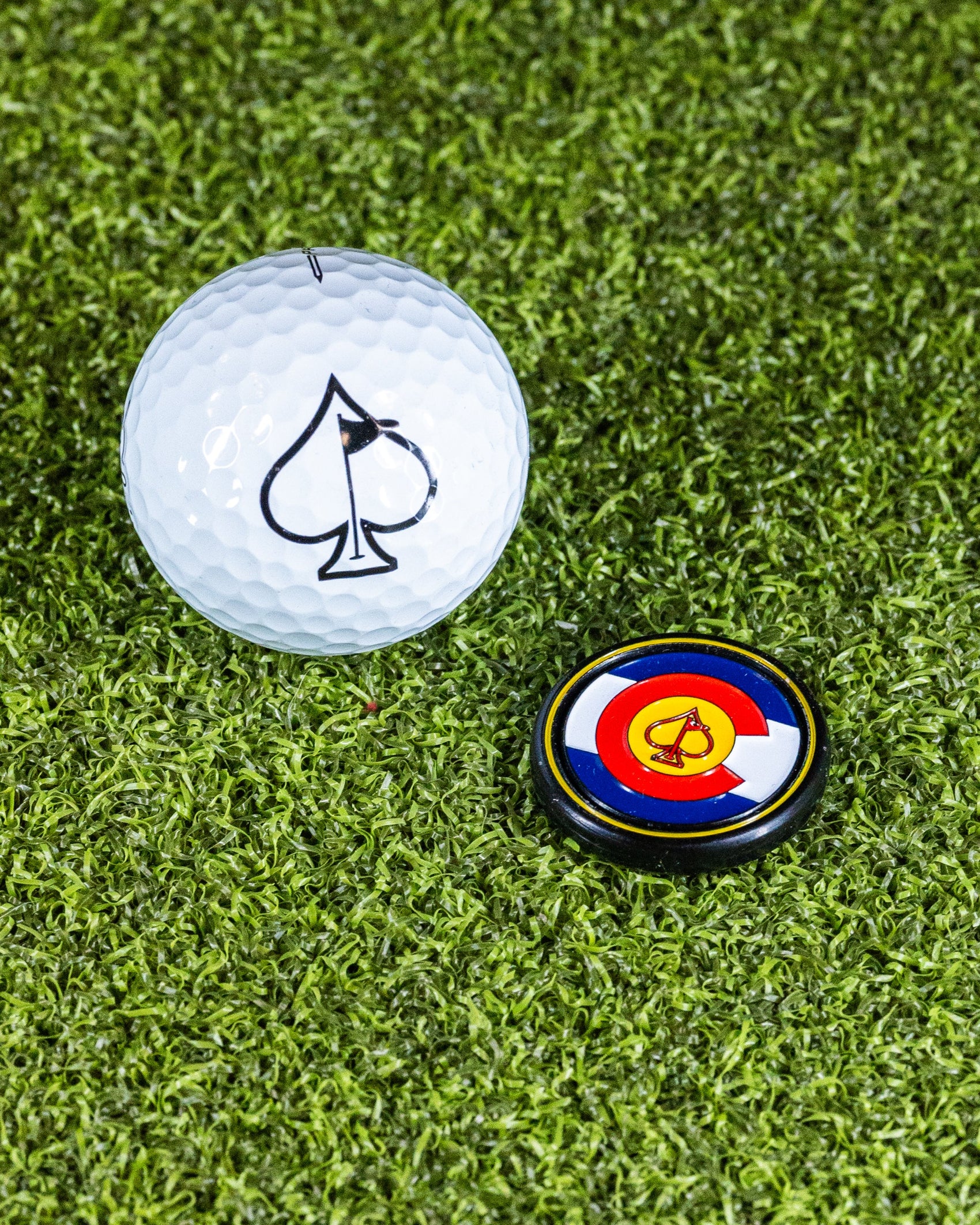 A white golf ball with a black spade symbol sits next to the Pins and Aces Pop Out Medallion Ball Marker - Colorado, both resting on green artificial turf.