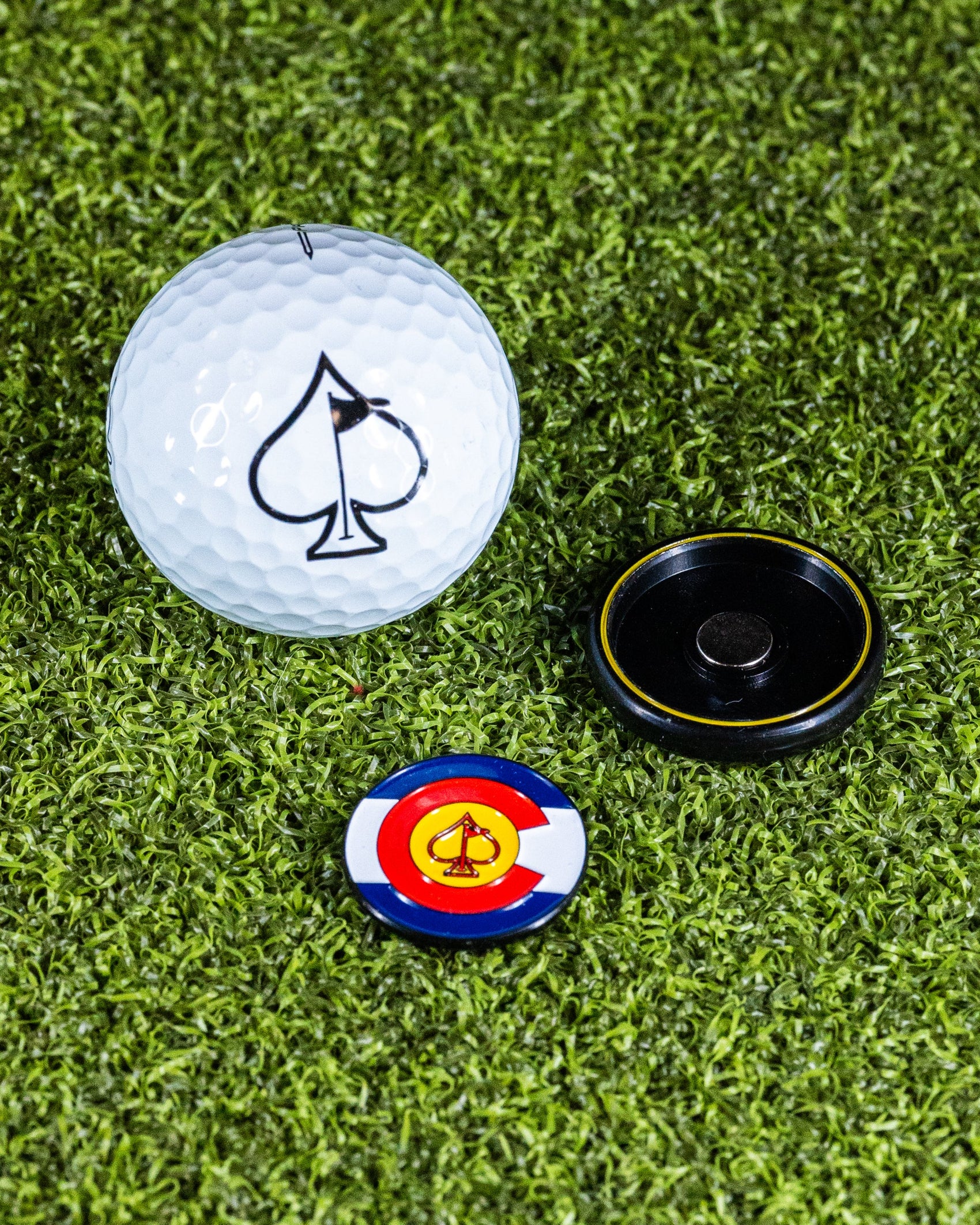 The Pins and Aces Pop Out Medallion Ball Marker - Colorado, featuring a spade and Colorado flag, comes with a black magnetic holder and golf ball, all displayed on green artificial turf.