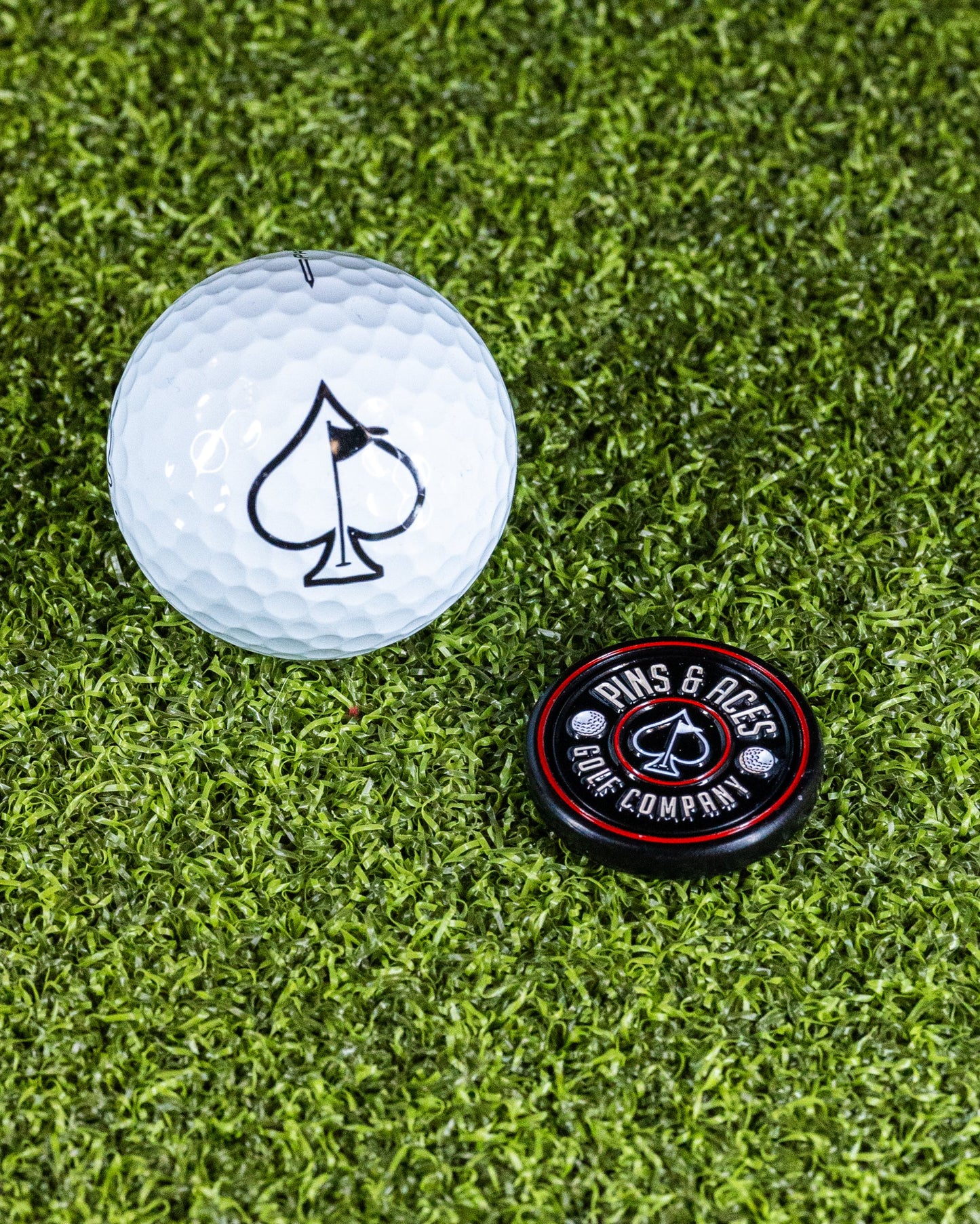 A Pop Out Medallion Ball Marker - Black by Pins and Aces with a spade logo lies on artificial grass beside a golf ball marked with a black spade.