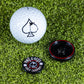 A white golf ball, the Pop Out Medallion Ball Marker - Black by Pins and Aces, and a circular marker are displayed on green artificial grass.