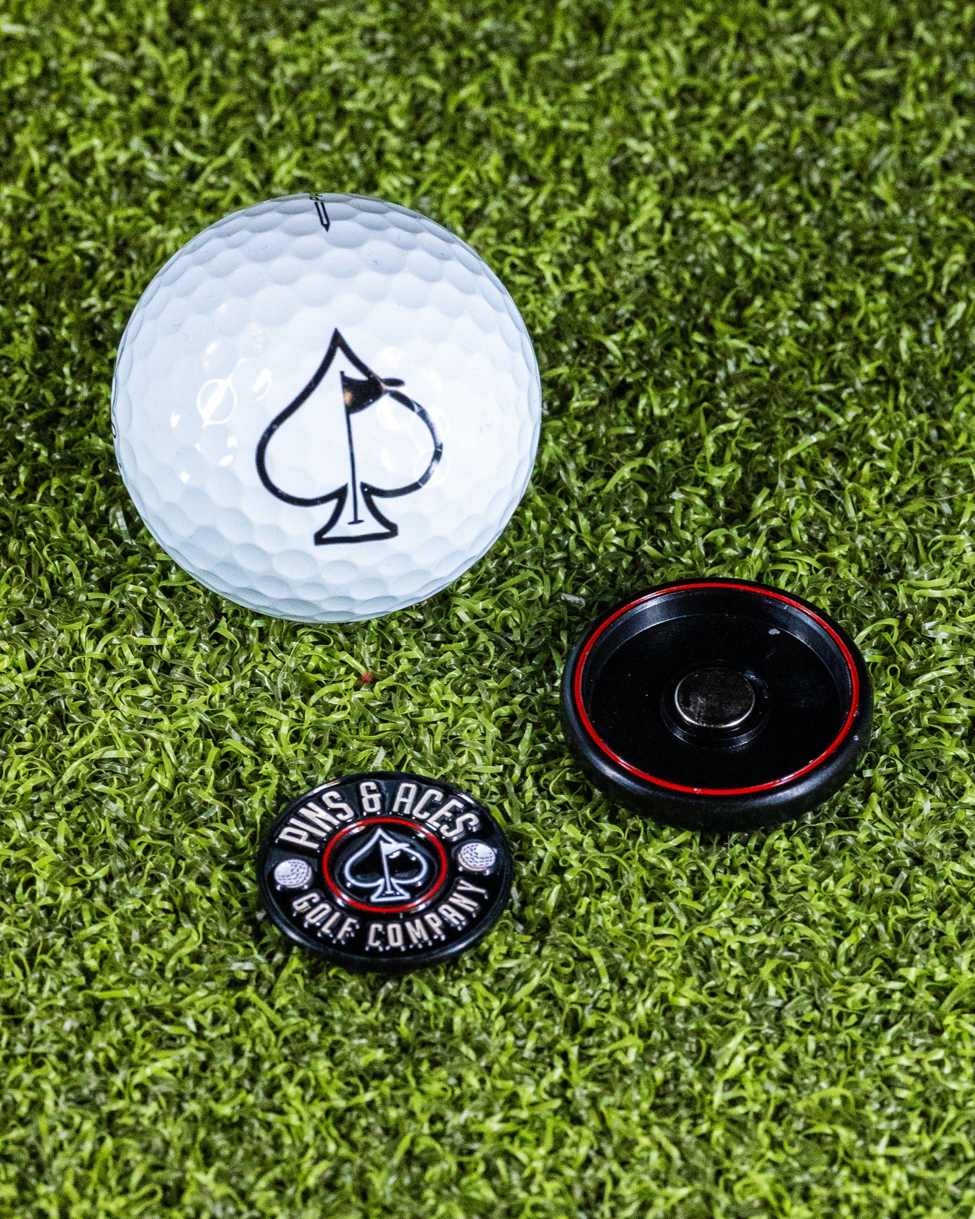 A white golf ball, the Pop Out Medallion Ball Marker - Black by Pins and Aces, and a circular marker are displayed on green artificial grass.