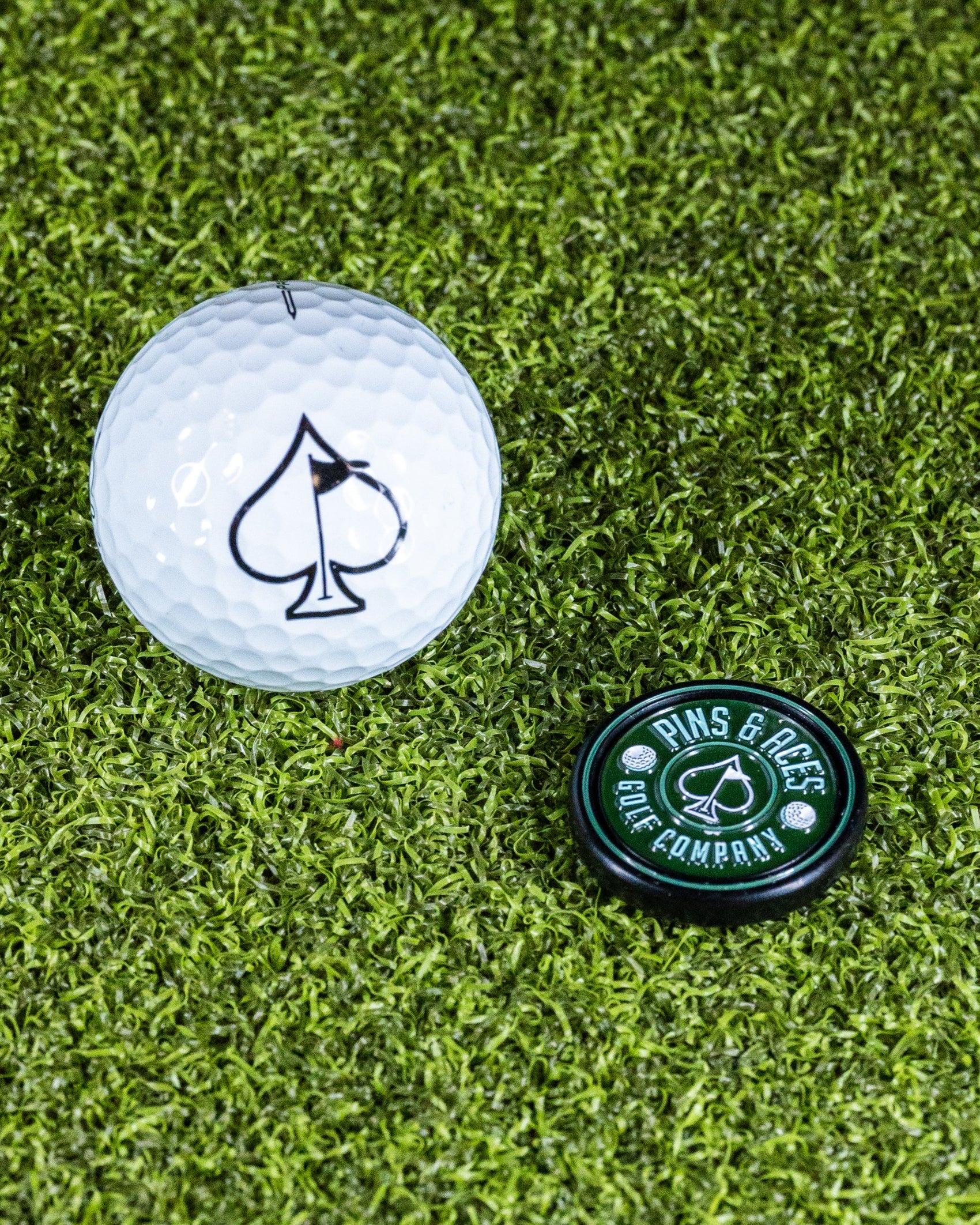 The Pop Out Medallion Ball Marker - Green by Pins and Aces features a magnetic ball marker with a black spade symbol, resting on bright green artificial grass—the ideal golf accessory for your next round.
