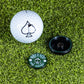 Pins and Aces’ Pop Out Medallion Ball Marker - Green, featuring a black spade symbol and a black holder, rests on green artificial grass—an essential golf accessory.