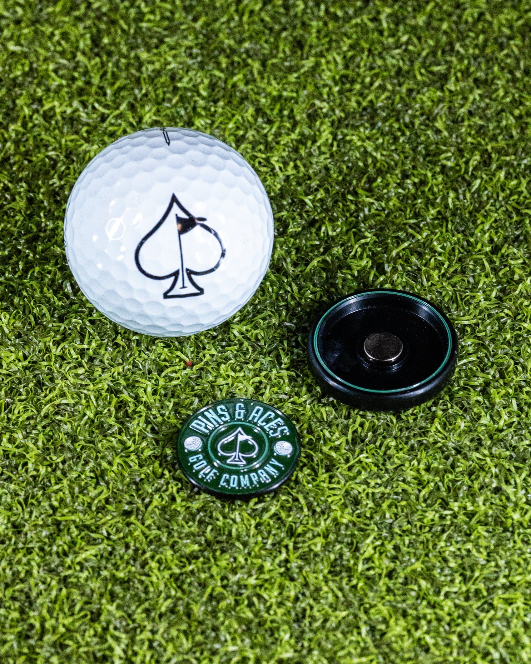 Pins and Aces’ Pop Out Medallion Ball Marker - Green, featuring a black spade symbol and a black holder, rests on green artificial grass—an essential golf accessory.
