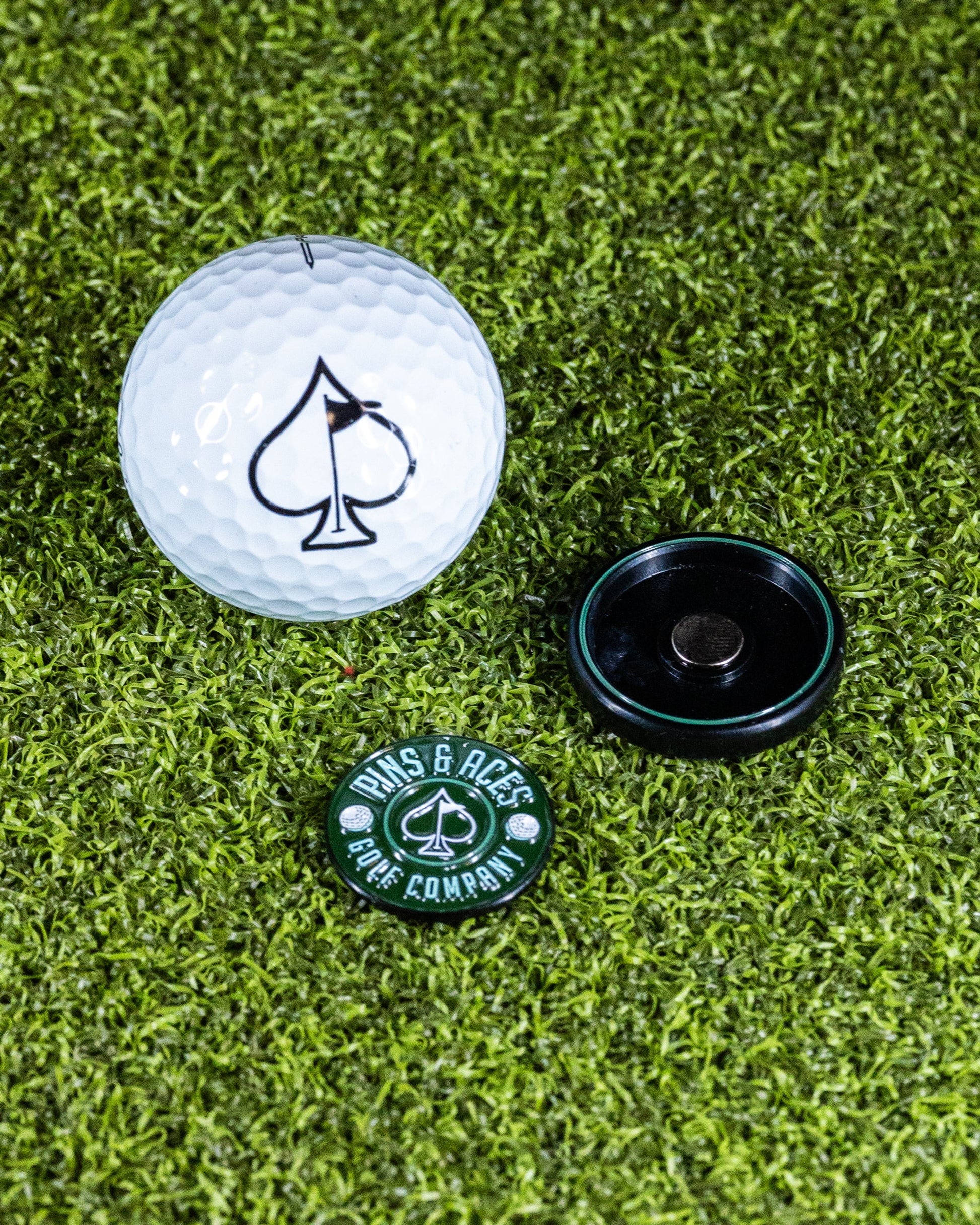 Pins and Aces’ Pop Out Medallion Ball Marker - Green, featuring a black spade symbol and a black holder, rests on green artificial grass—an essential golf accessory.