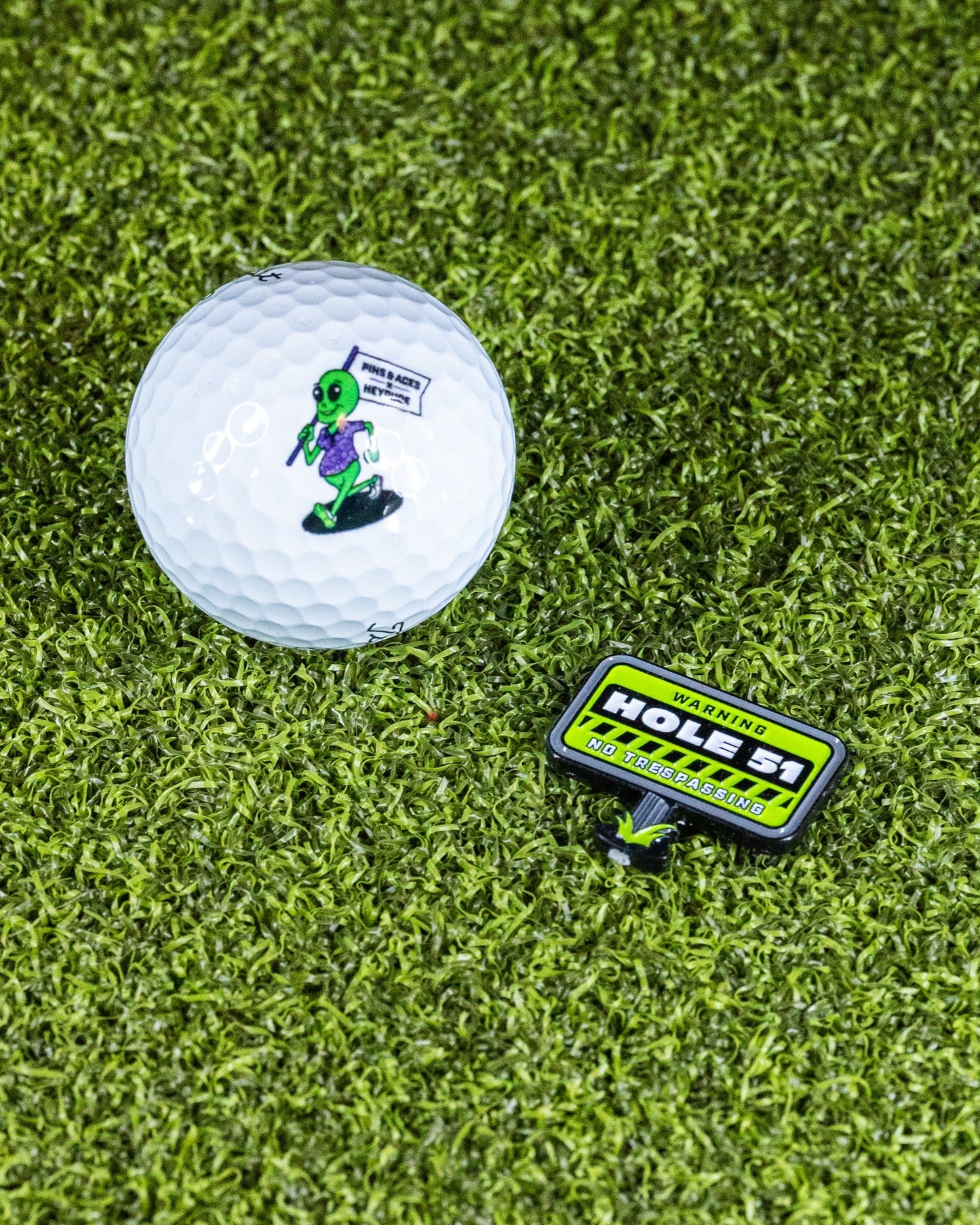 A golf ball with a green alien holding a flag that says "TAKE ME TO YOUR GOLF COURSE," paired with the Pins and Aces HEYDUDE - Hole 51 Ball Marker, which reads: WARNING: HOLES! NO TRESPASSING, set on artificial grass.