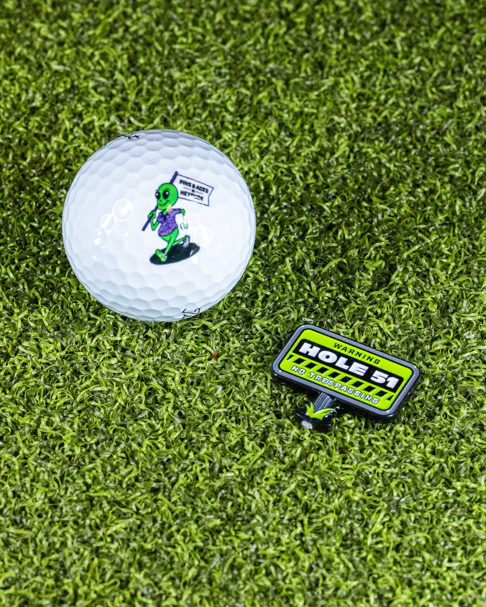 A golf ball with a green alien holding a flag that says "TAKE ME TO YOUR GOLF COURSE," paired with the Pins and Aces HEYDUDE - Hole 51 Ball Marker, which reads: WARNING: HOLES! NO TRESPASSING, set on artificial grass.