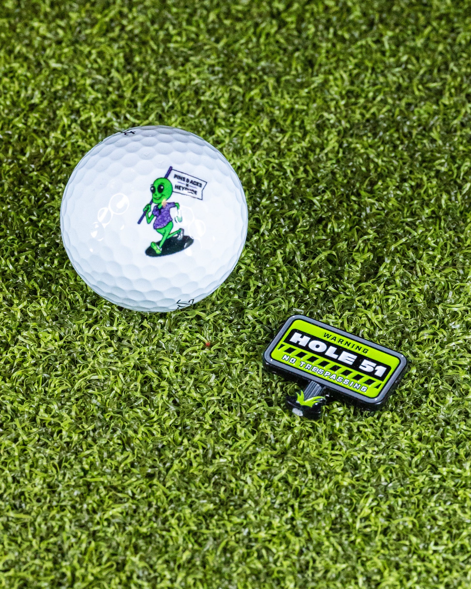 A golf ball with a green alien holding a flag that says "TAKE ME TO YOUR GOLF COURSE," paired with the Pins and Aces HEYDUDE - Hole 51 Ball Marker, which reads: WARNING: HOLES! NO TRESPASSING, set on artificial grass.