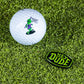 A golf ball on grass rests beside a sleek Pins and Aces HEYDUDE Logo Ball Marker, ready for your next putt.