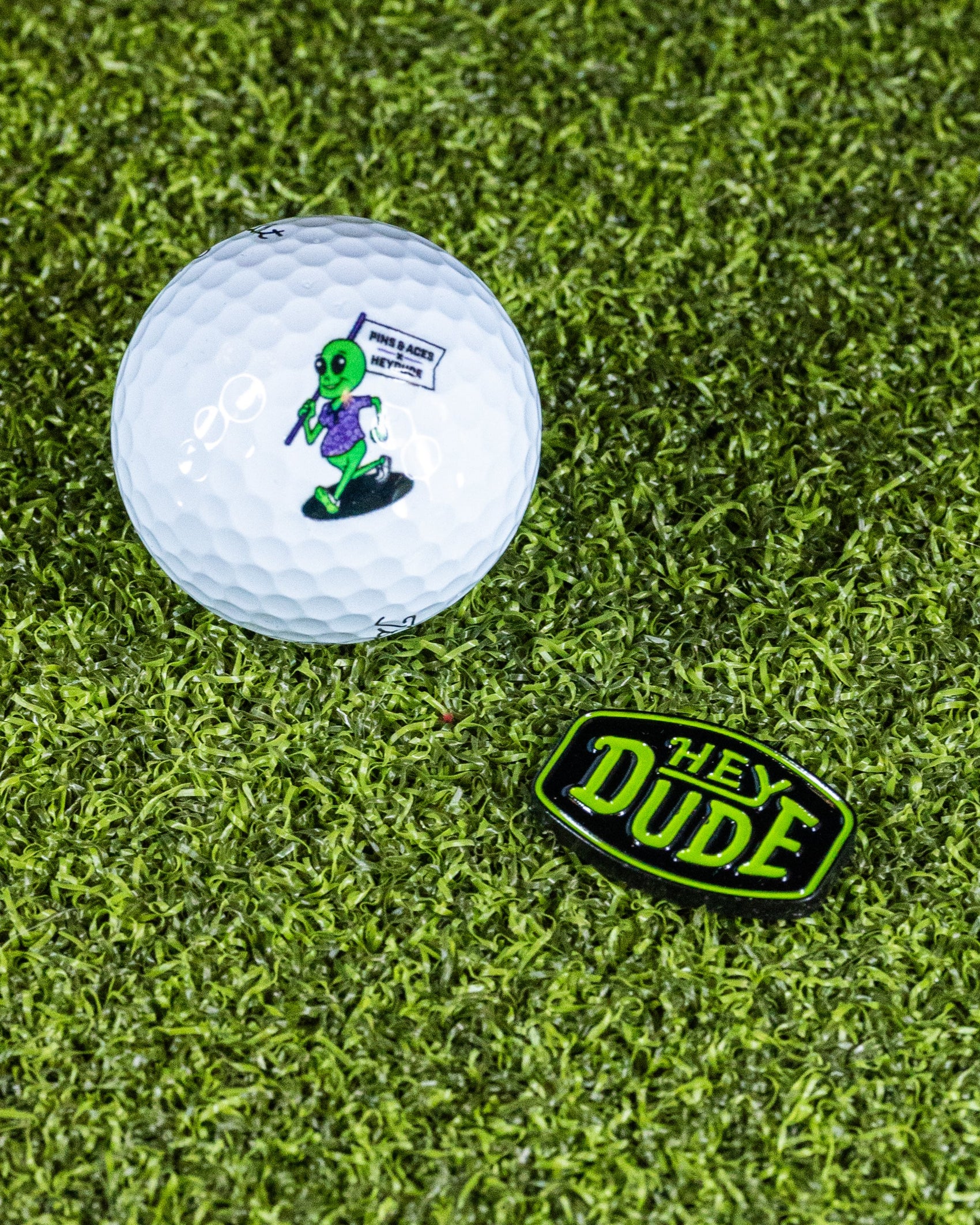 A golf ball on grass rests beside a sleek Pins and Aces HEYDUDE Logo Ball Marker, ready for your next putt.