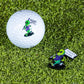 A Pins and Aces HEYDUDE - Alien Ball Marker and matching golf ball, both featuring the alien design with a "Never Judge" flag, are placed on green grass.