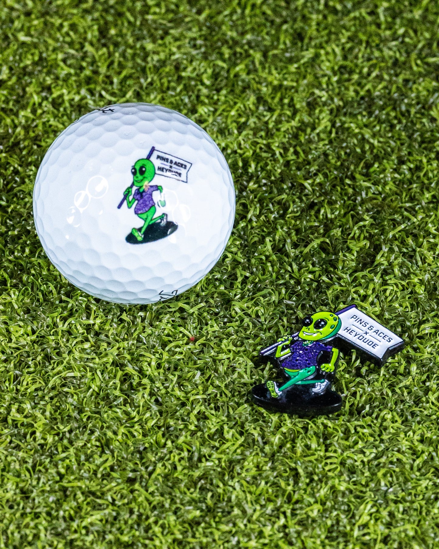 A Pins and Aces HEYDUDE - Alien Ball Marker and matching golf ball, both featuring the alien design with a "Never Judge" flag, are placed on green grass.