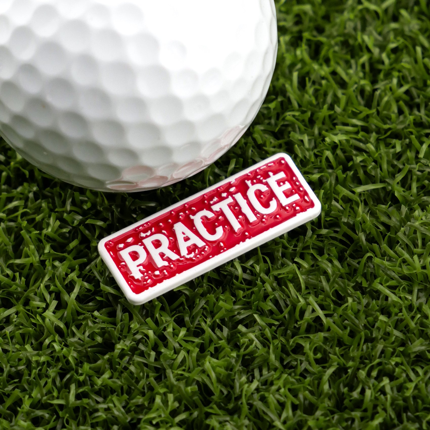 A close-up of a golf ball on artificial turf next to Logie Joe's Golf Practice Stamp, a red and white badge that boldly displays “PRACTICE”—a premium accessory for marking your balls on the green.