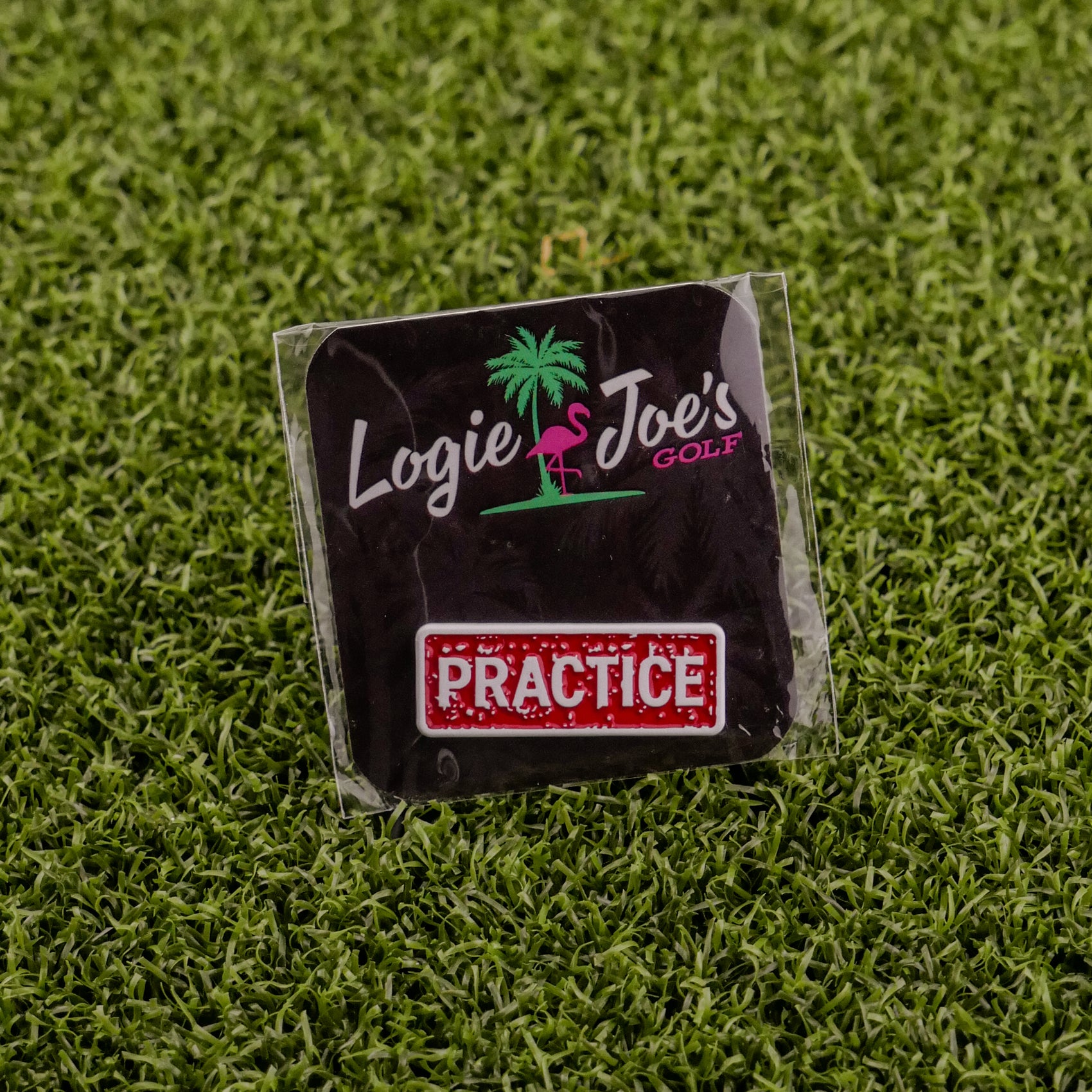 The Practice Stamp by Logie Joe's Golf features a pink flamingo logo and red PRACTICE sign on a sleek black square, displayed on green artificial grass—a premium accessory for golf enthusiasts.