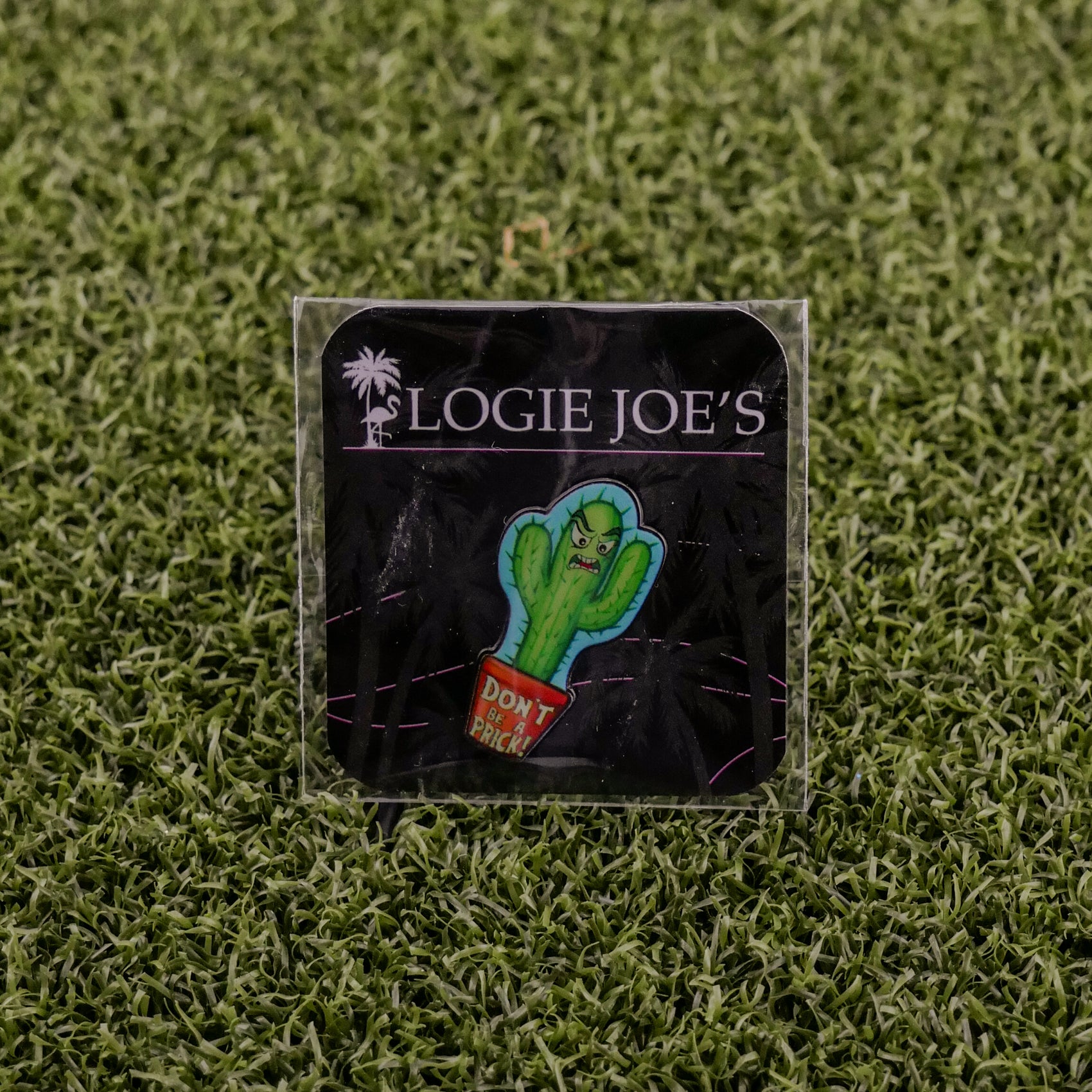 The Don’t be a Prick cactus pin by Ballmarkers by Logie Joe’s, featuring an angry face and red pot on branded black backing over faux grass, is ideal for anyone seeking a funny golf accessory or unique golf ball marker.