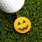 The Pumpkin And Tee Ball Marker by Ballmarkers.com features a jack-o-lantern face and dimpled texture, adding Halloween fun to your golf game. Shown on green grass, it’s a playful take on traditional golf accessories.