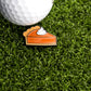 A golf ball rests on artificial grass next to the Pumpkin Pie Ball Marker by Ballmarkers.com—a small enamel pin shaped like a slice of pumpkin pie with whipped cream—adding a fun reminder that there’s always time to celebrate dessert.