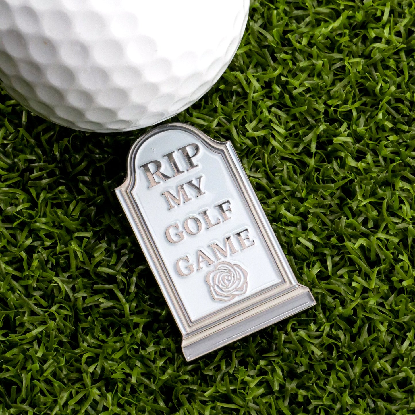 The RIP My Golf Game Tombstone Ball Marker by Ballmarkers.com, engraved with "RIP MY GOLF GAME" and a rose, rests beside a golf ball on artificial grass—an unforgettable accessory for any golfer.