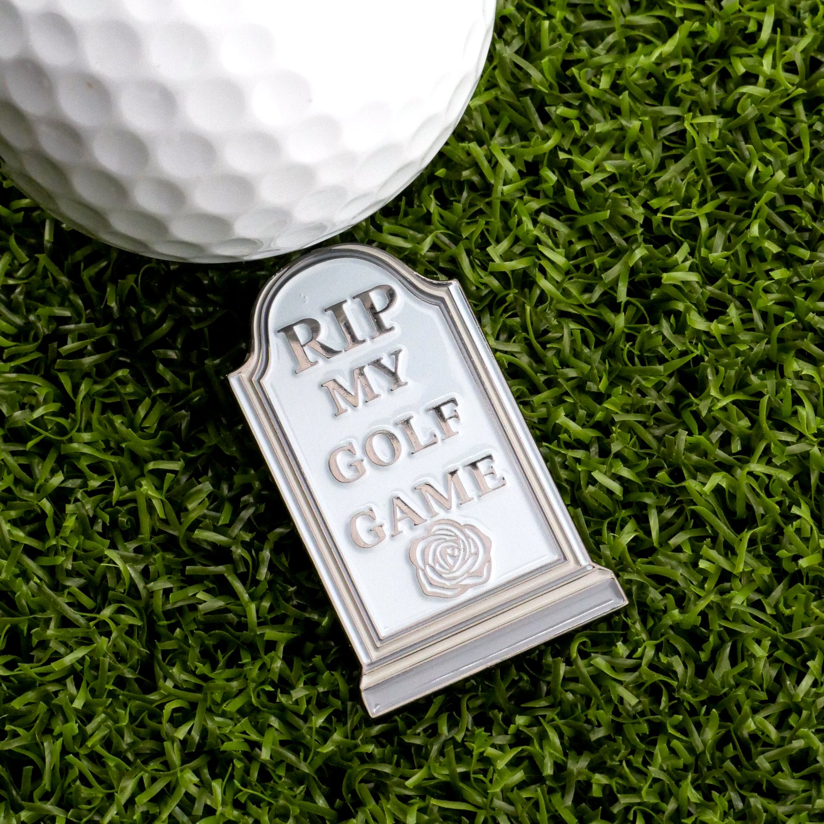 The RIP My Golf Game Tombstone Ball Marker by Ballmarkers.com, engraved with "RIP MY GOLF GAME" and a rose, rests beside a golf ball on artificial grass—an unforgettable accessory for any golfer.