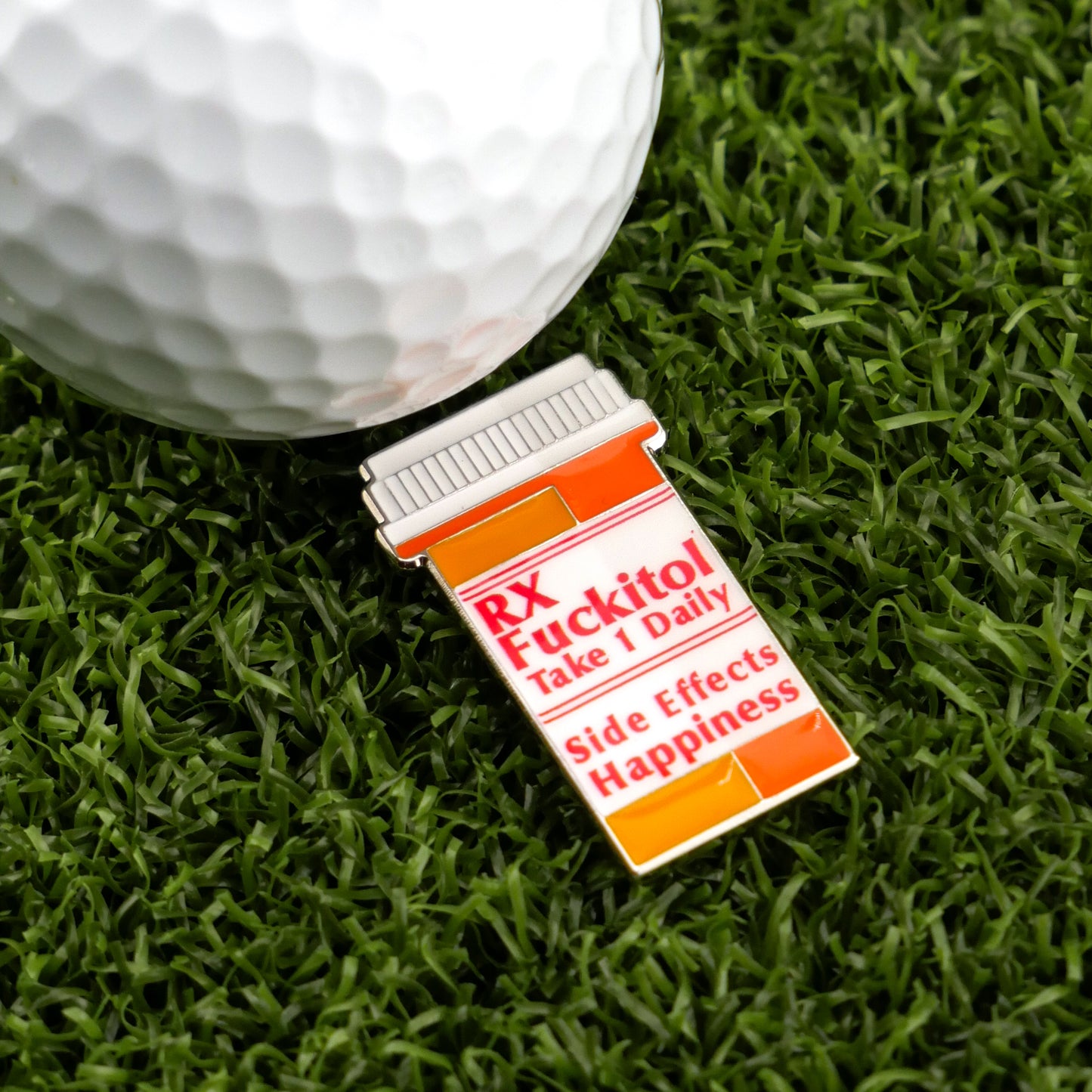 The Logie Joe's Golf "RX Fuckitol" novelty pin, shaped like a pill bottle and labeled “Take 1 Daily. Side Effects: Happiness,” adds a quirky touch to your golf accessories—shown here beside a golf ball on artificial grass.