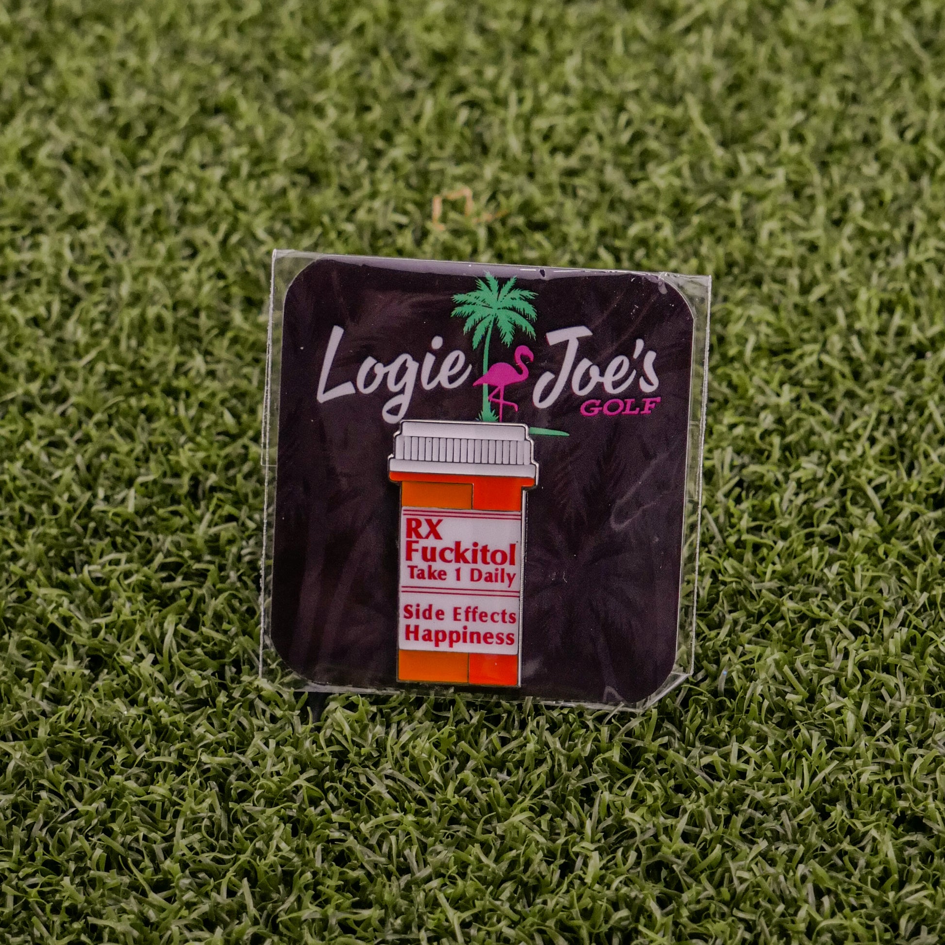 A square sticker on grass displays Ballmarkers by Logie Joe’s RX Fuckitol above a cartoon pill bottle labeled Take 1 Daily. Side effects: happiness, palm tree & flamingo—great for fans of funny golf accessories or unique golf ball markers.