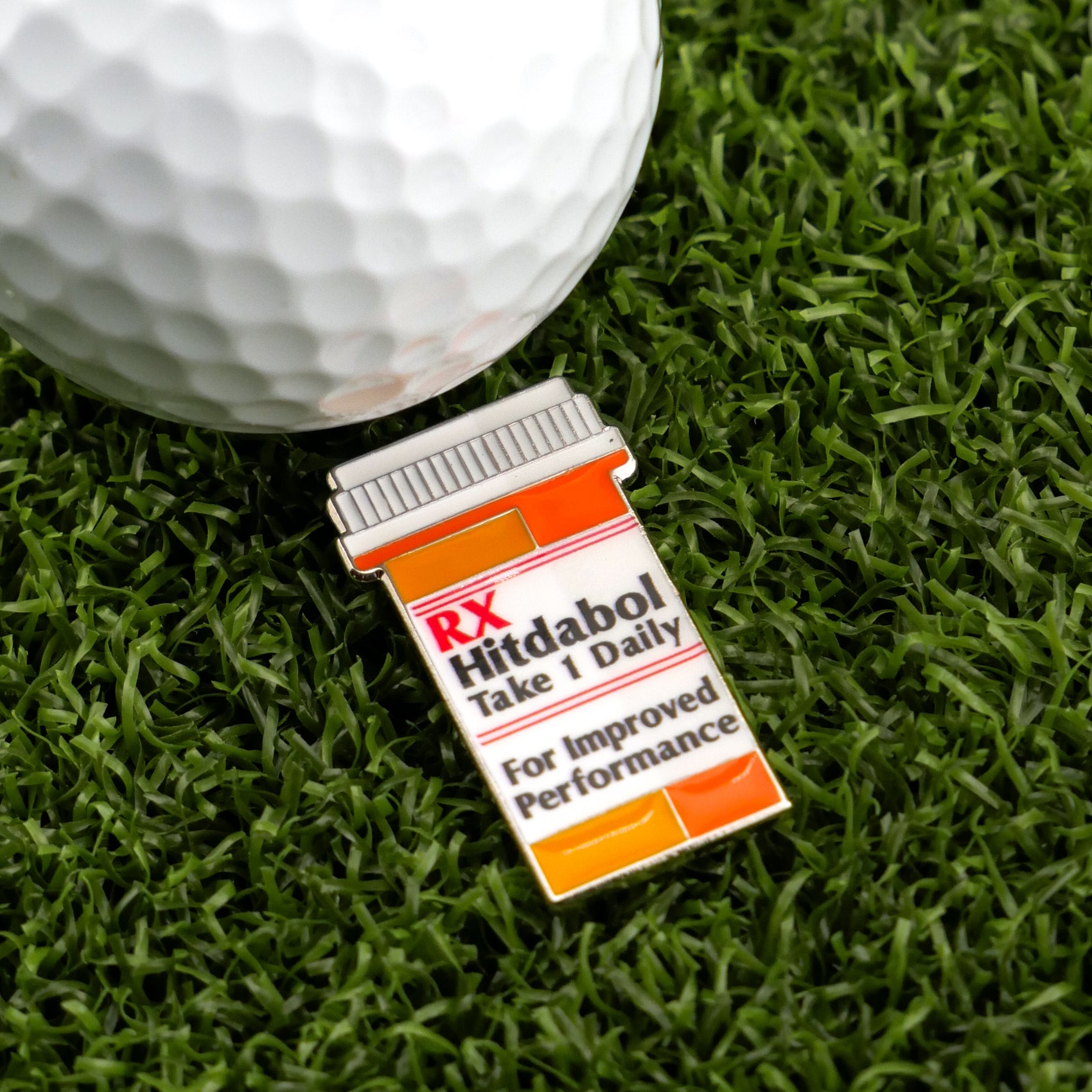 A golf ball rests on green grass beside a Ballmarkers.com premium marker shaped like a prescription bottle labeled RX Hitdabol.