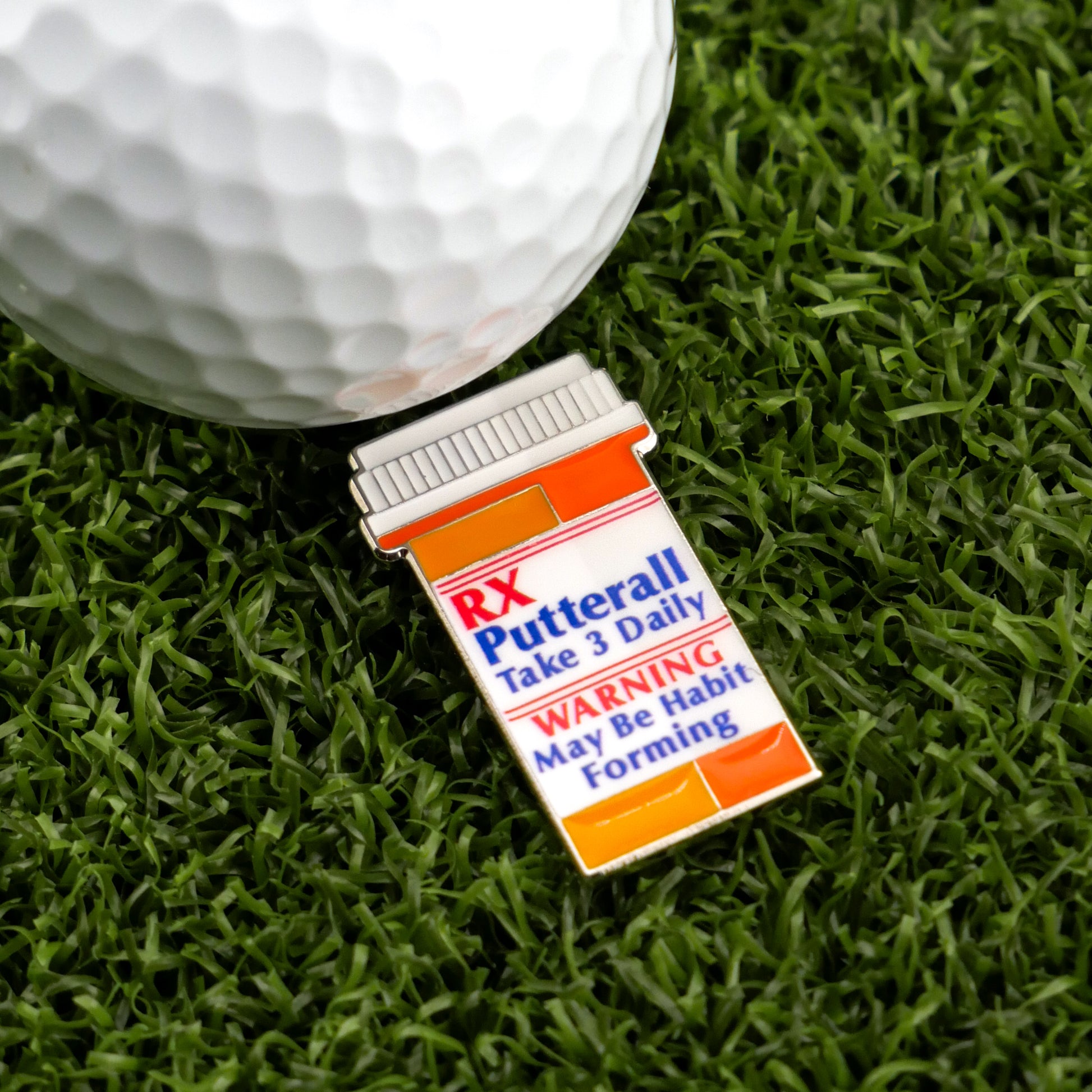 A golf ball on artificial grass sits beside Logie Joe's Golf RX Putterall—a novelty pin shaped like a prescription bottle labeled “Take 3 Daily; WARNING May Be Habit Forming”—and a magnetic ball marker for your next putt.