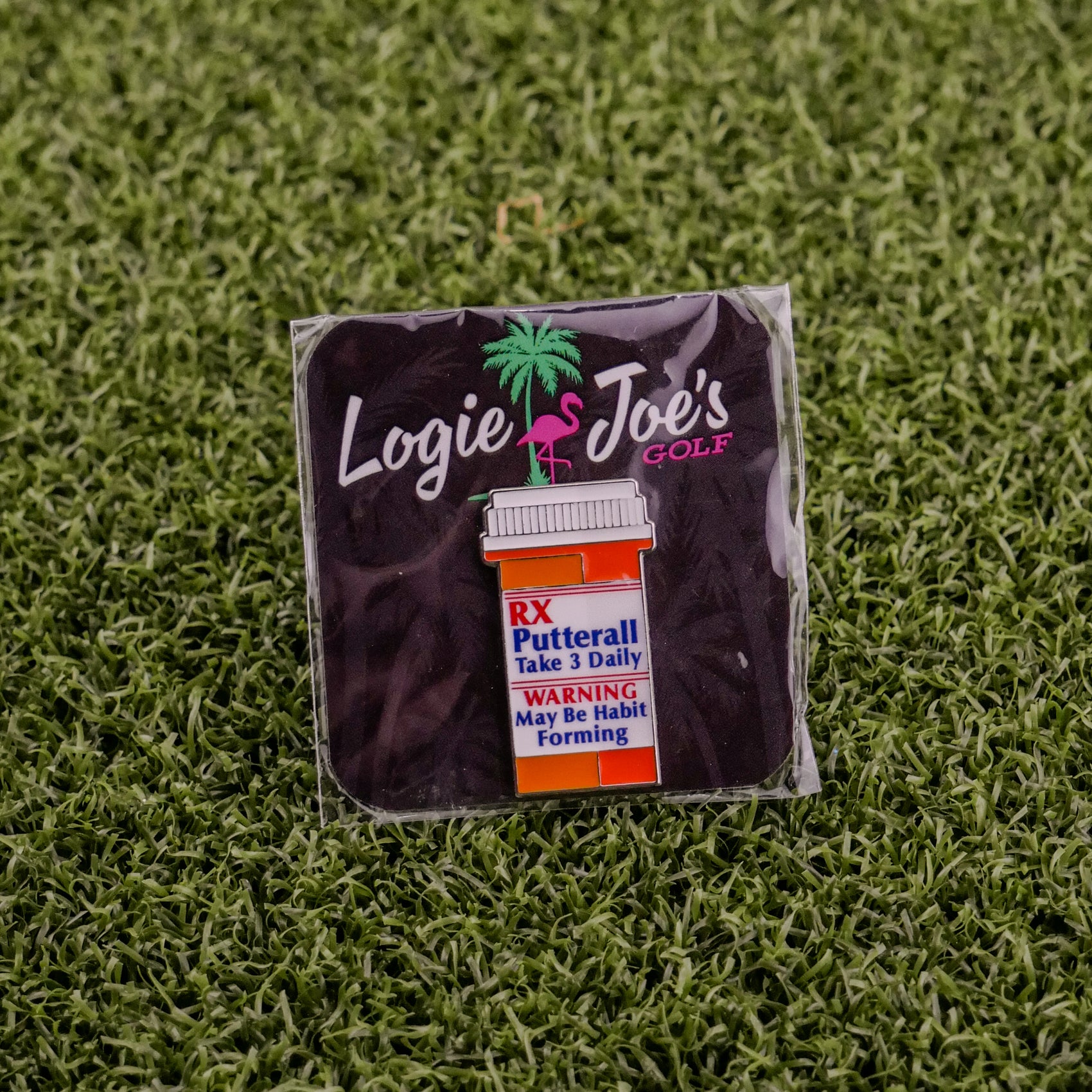 The RX Putterall is a golf ball marker shaped like a prescription pill bottle, labeled “Take 3 Daily.” Set on artificial grass with Logie Joe’s Golf branding, it’s a unique accessory for any golfer.