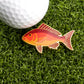 The Logie Joe's Golf Red Snapper Ball Marker, shaped like a red and yellow fish, sits on green grass beside a white golf ball, bringing playful style to premium golf accessories.