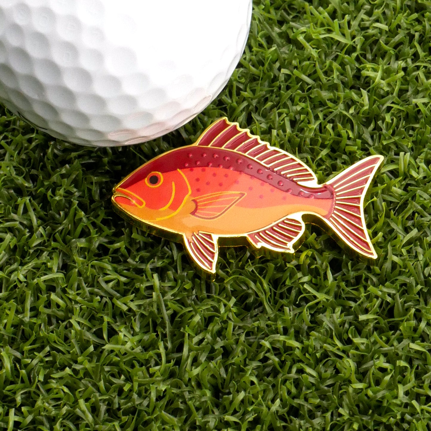 The Logie Joe's Golf Red Snapper Ball Marker, shaped like a red and yellow fish, sits on green grass beside a white golf ball, bringing playful style to premium golf accessories.
