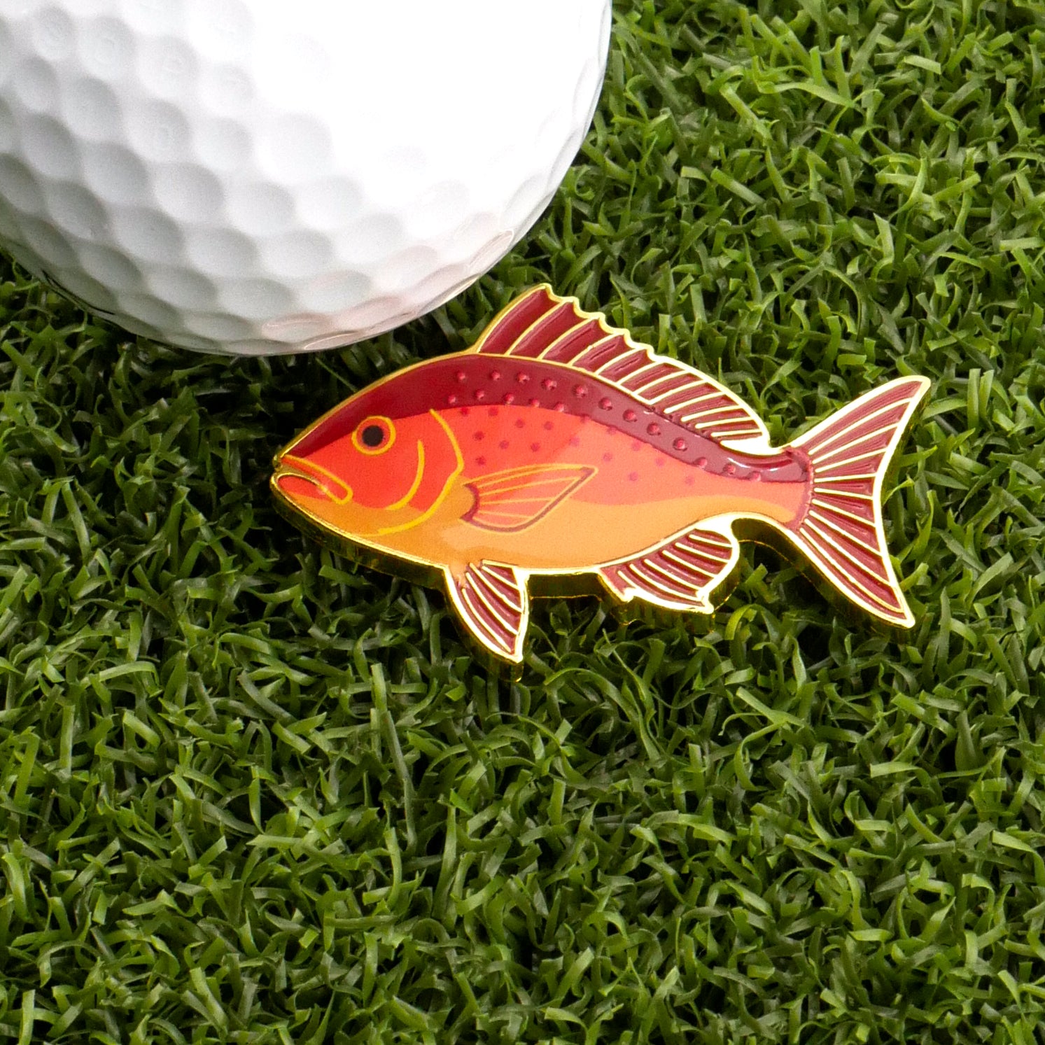 The Logie Joe's Golf Red Snapper Ball Marker, shaped like a red and yellow fish, sits on green grass beside a white golf ball, bringing playful style to premium golf accessories.
