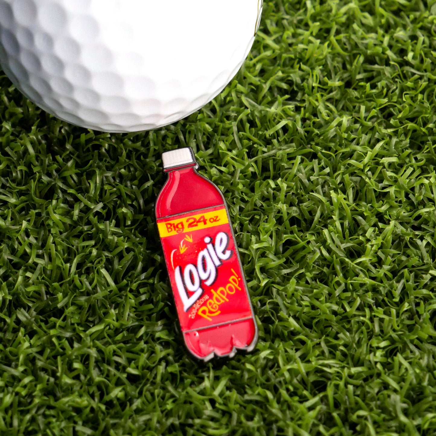 A 24-ounce red Logie Joe's Golf Redpop Ball Marker, inspired by classic soda, sits on artificial grass beside a white golf ball—ideal as a fun and unique golf accessory.