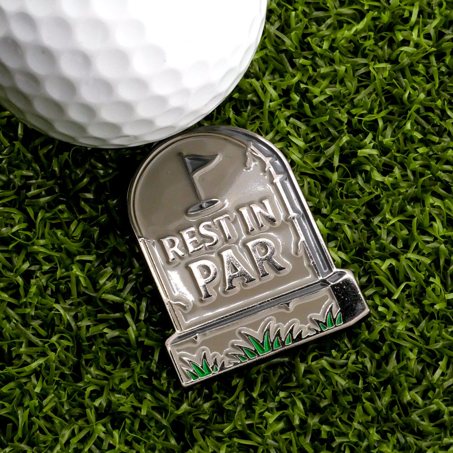 The Rest In Par Ball Marker by Ballmarkers.com is a metal tombstone-shaped golf accessory featuring a golf flag design and the words "Rest in Par," shown on green artificial grass beside a white golf ball.